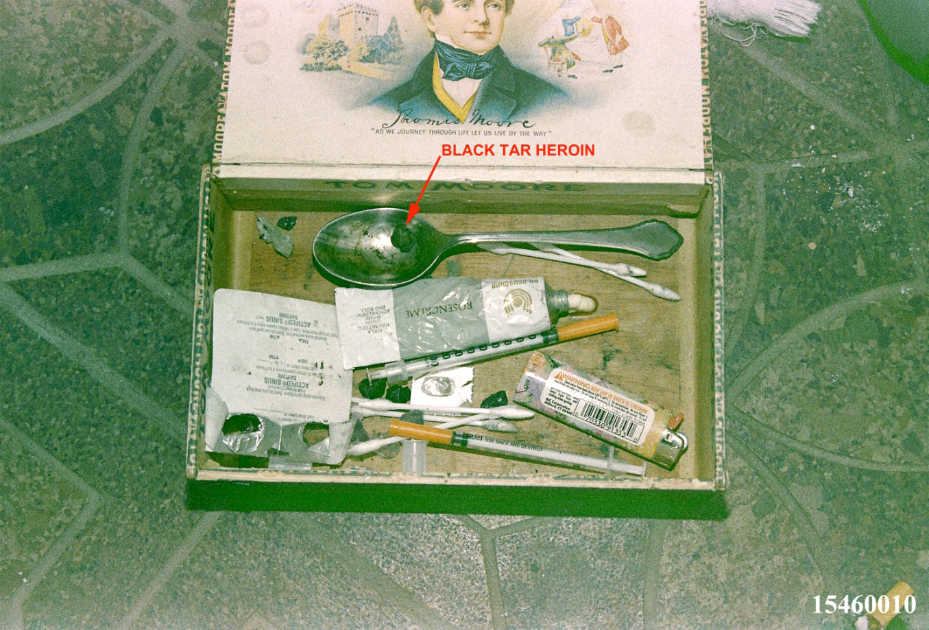Figure 16: Cobain’s heroin prep cigar box found at the scene; the two syringes were the insulin type with the hub as part of the syringe. Of these two insulin syringes, one was speculated by the police investigators to have been used to inject the lethal heroin dose before the shotgun discharge [2]. These syringes appear to be from the same manufacturer as the one in Figure 14A2. There is a black tar heroin rock on the spoon and others in the box.